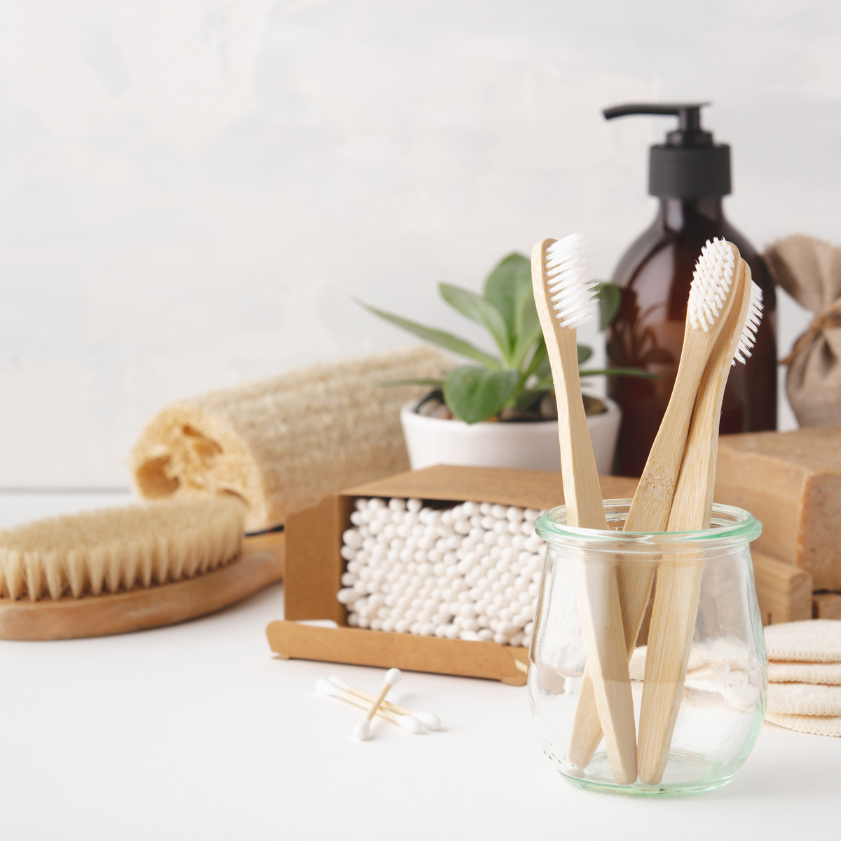 3 bamboo sustainable toothbrushes standing up in a jar, with eco-friendly cotton swabs and hairbrush in the back ground.  A small plant is next to a loofah sponge