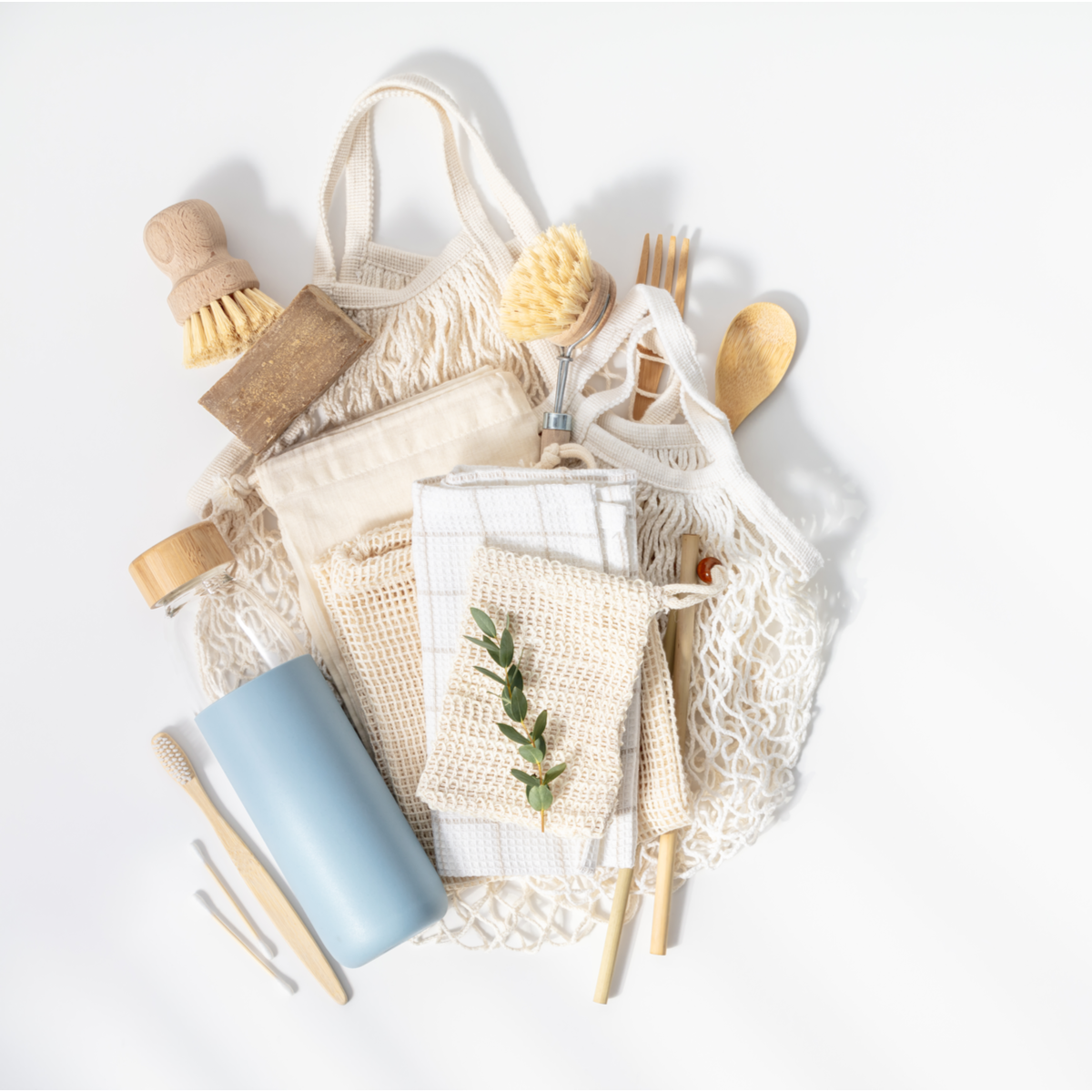 Various eco-friendly products in a pile, including cotton and muslin soap and grocery bags, sisal scrubbing brush and a reusable blue drinking mug
